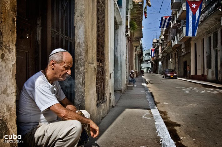 Old jewish man sitting on the street in the left and a cuban flag in the back on the right © Cuba Absolutely, 2014